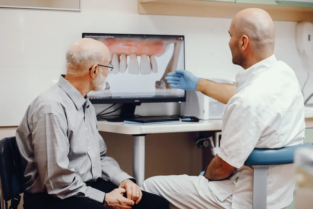 A dentist explains a dental procedure to an elderly patient using a computer screen, emphasizing how to reduce dental expenses with the Rubicare Dental Savings Plan.